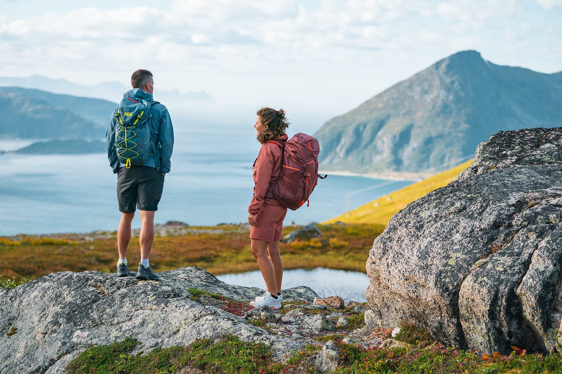 Two hikers with backpacks stand on rocky terrain, overlooking a scenic coastal landscape with mountains and ocean.