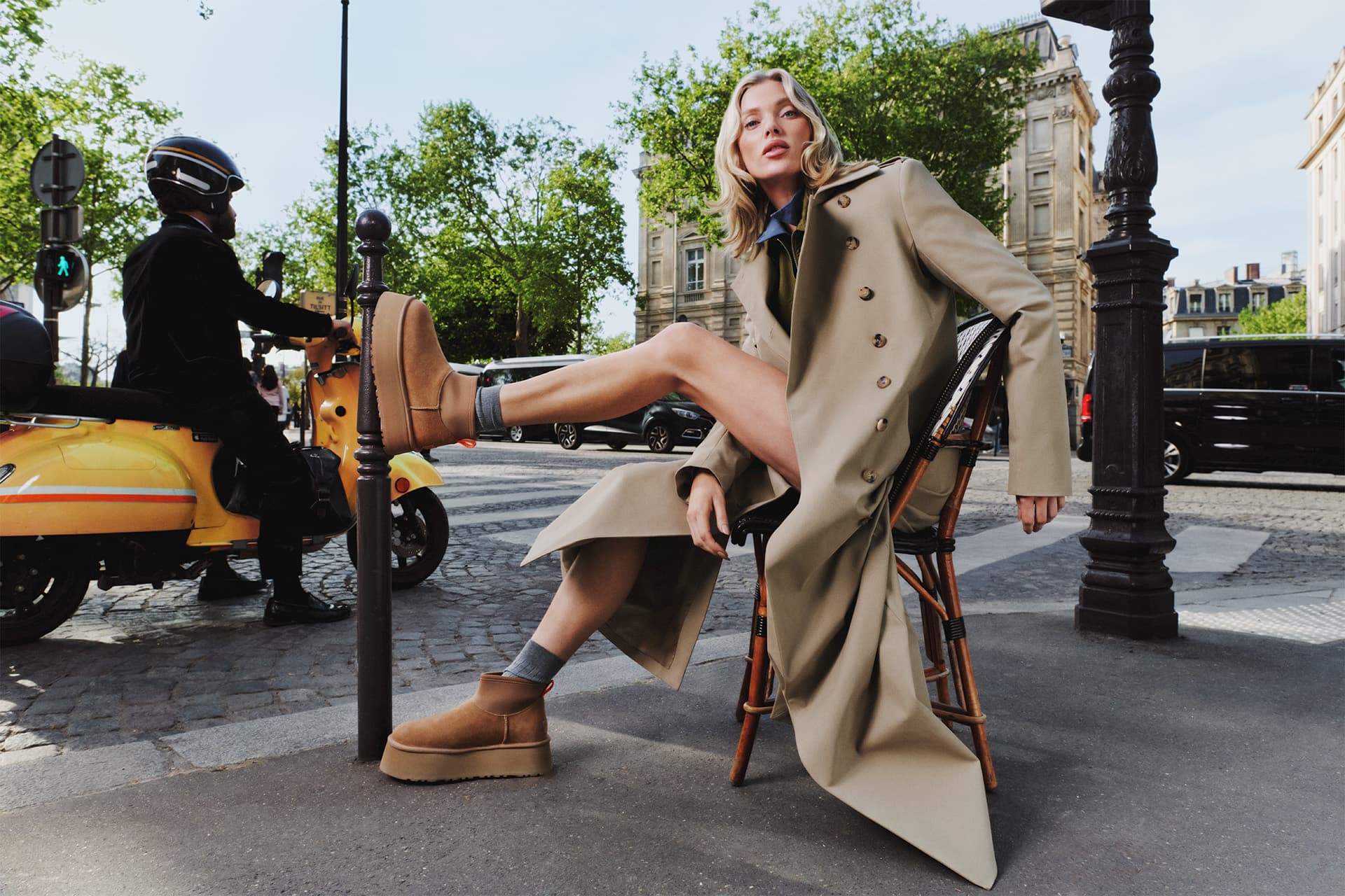 A person in a long beige coat poses confidently on a chair by the street, with one leg raised, wearing tan platform shoes.