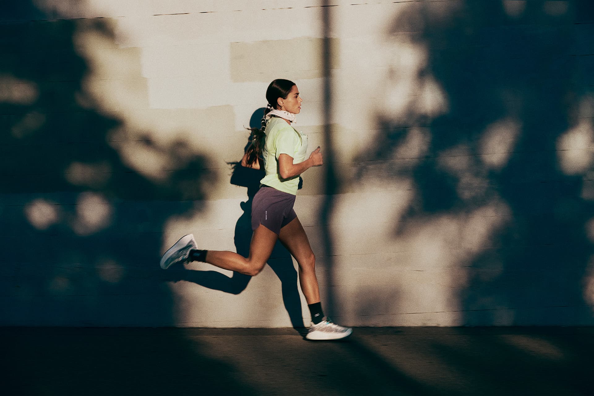 Woman jogging against a sunlit wall, casting a shadow. She wears a light shirt, shorts, and white sneakers. The background shows dappled shadows.