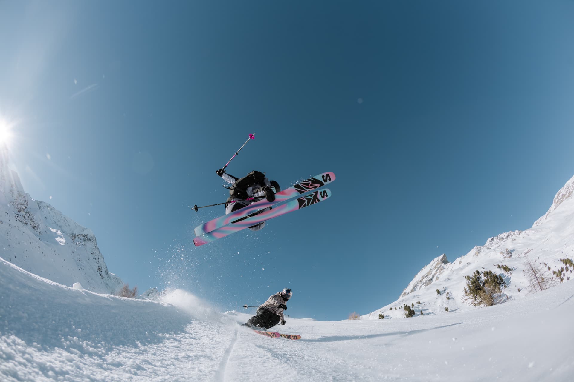 Two skiers performing jumps on a snowy slope under a clear blue sky, with one skier mid-air and the sun shining brightly.