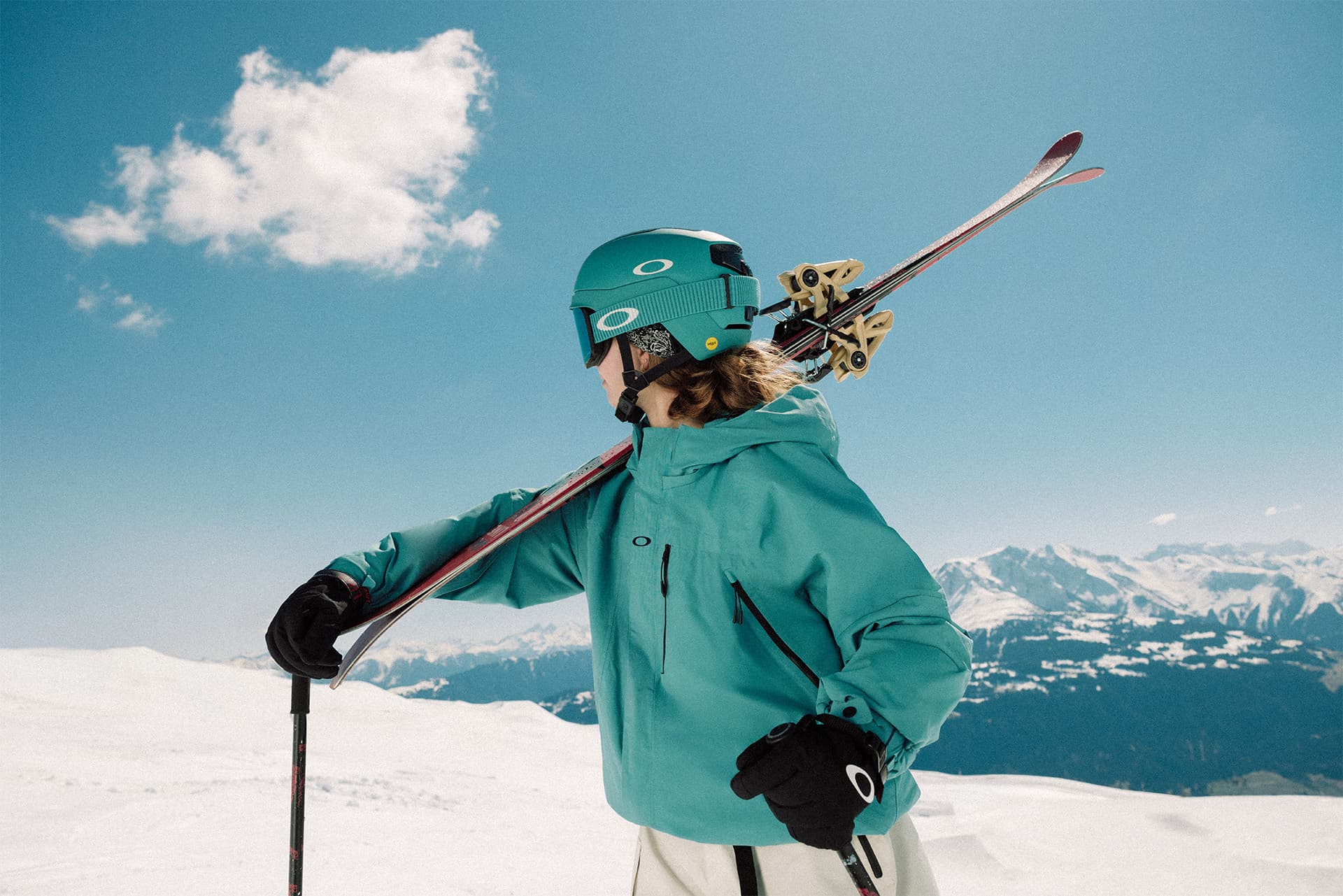 Skier in turquoise gear holds skis on shoulder, standing on a snowy slope with a clear blue sky and distant mountains.