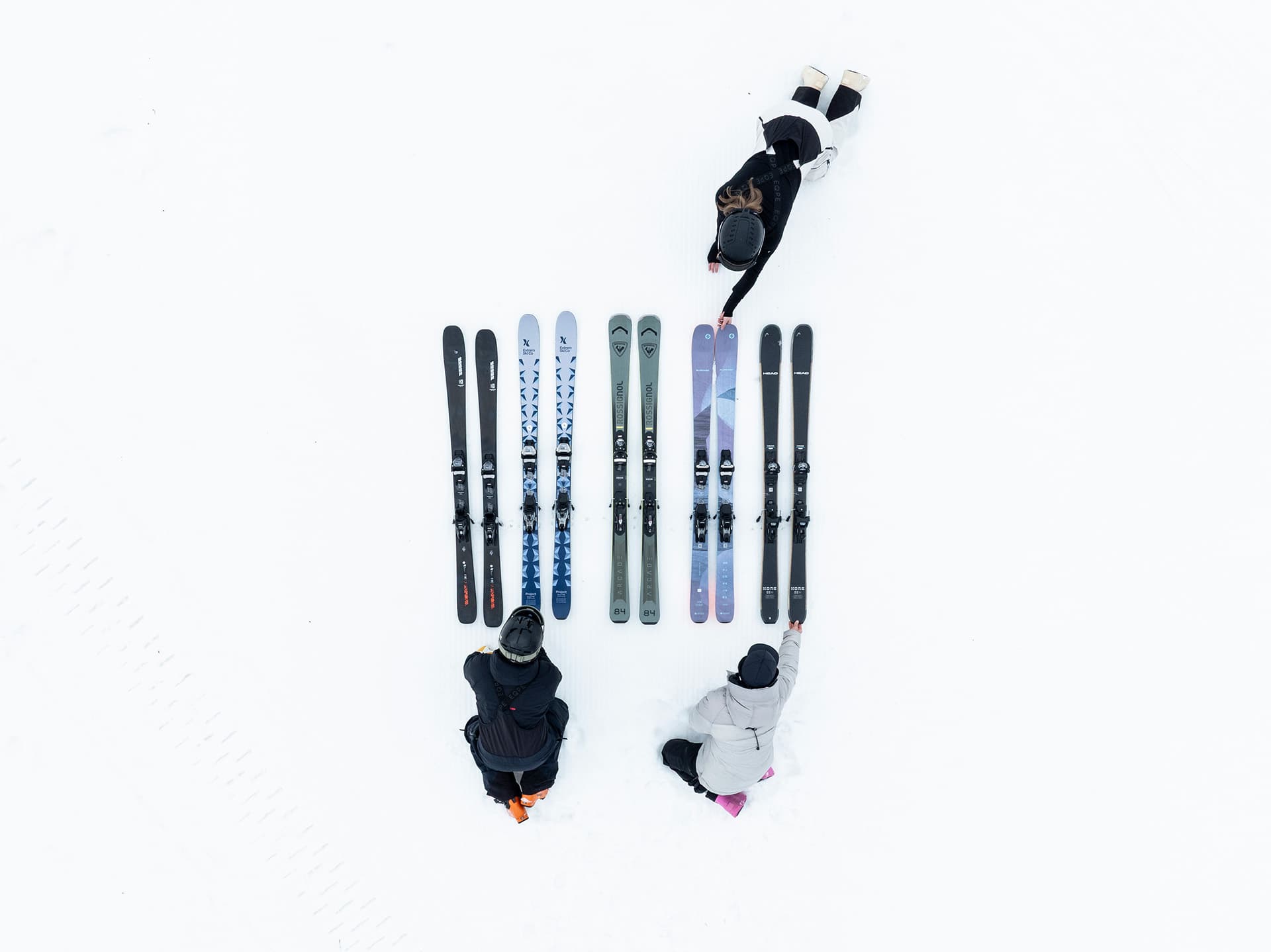 Shot from above of three people and five pair of skis laying flat against snowy ground
