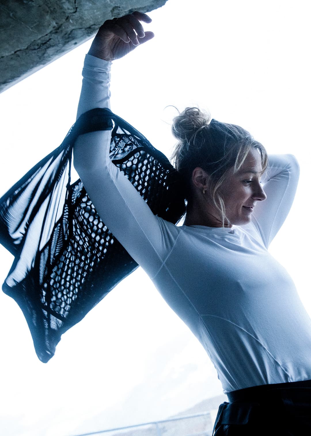 Woman in a white top holding a net-like black fabric over her shoulder, with her hair in a bun, against a bright background.