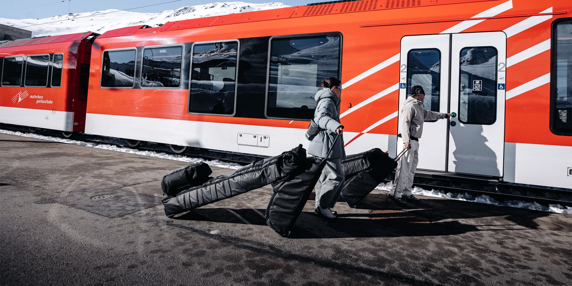 Two people in winter clothing board a red train, each pulling a large ski bag, with snow visible in the background.