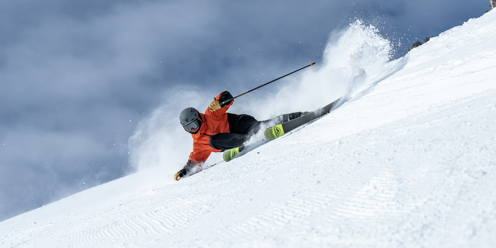 Skier in an orange jacket and black helmet carving sharply down a snowy slope, with snow spraying up behind against a cloudy sky.