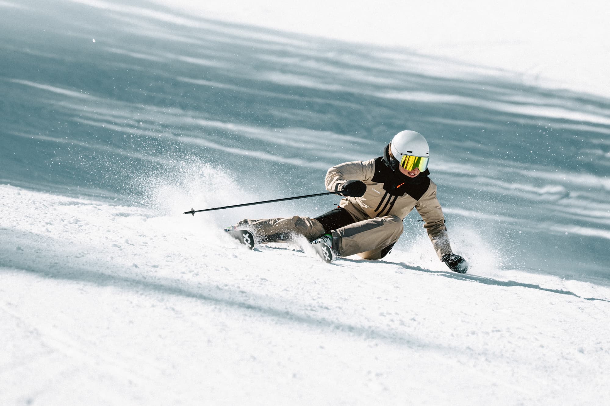 Skier in beige ski clothes carving through snow on a sunny slope, creating a spray of powder behind.