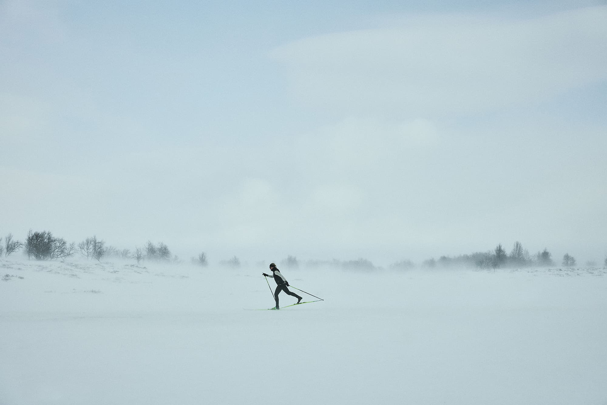 A person cross-country skiing through a vast, snowy landscape under a cloudy sky, with distant trees barely visible in the mist.