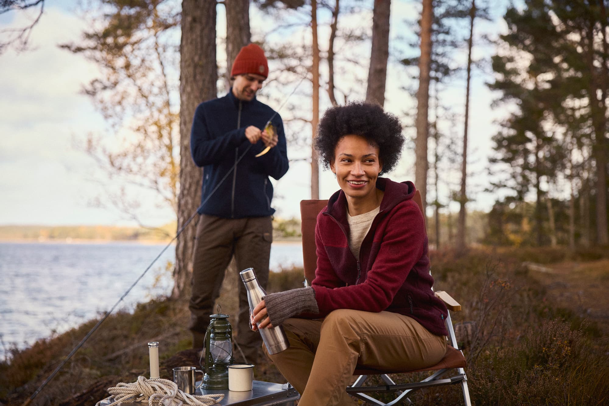 A woman sits smiling with a thermos near a lake, while a man, wearing a red beanie, prepares a fishing rod in a wooded area.