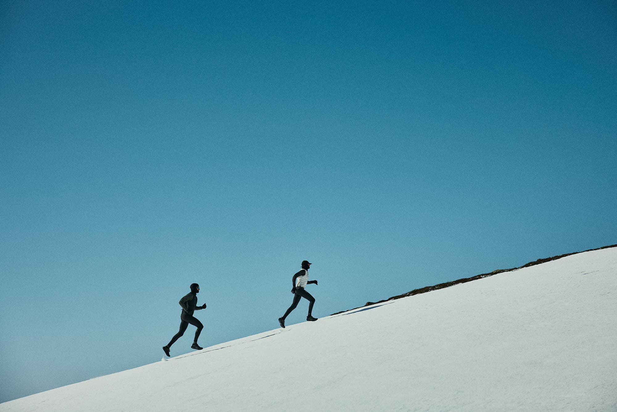 Two people running uphill on a snowy slope against a clear blue sky.