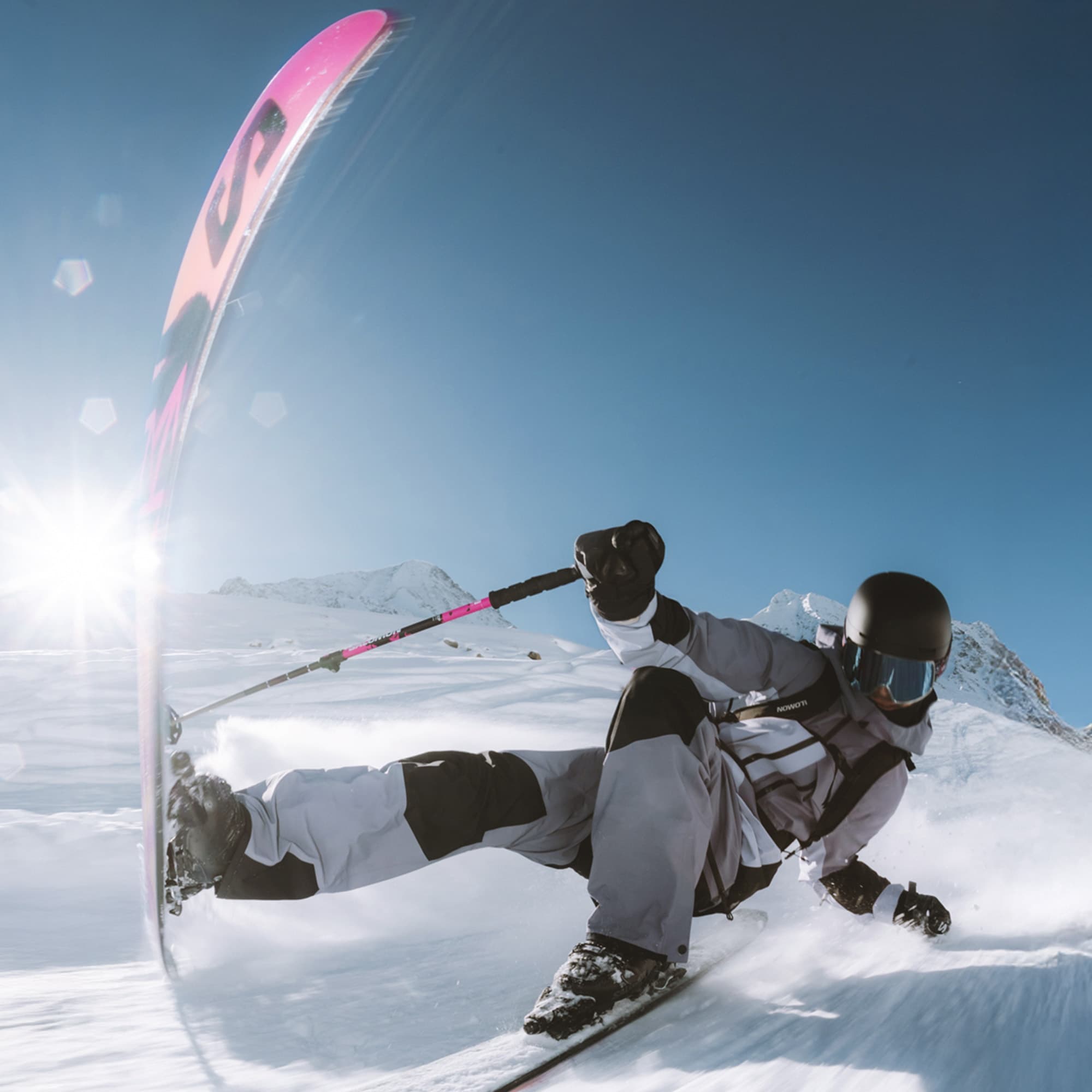 Skiier in action, carving through snow on a sunny day, with a bright sunburst and snowy mountains in the background.