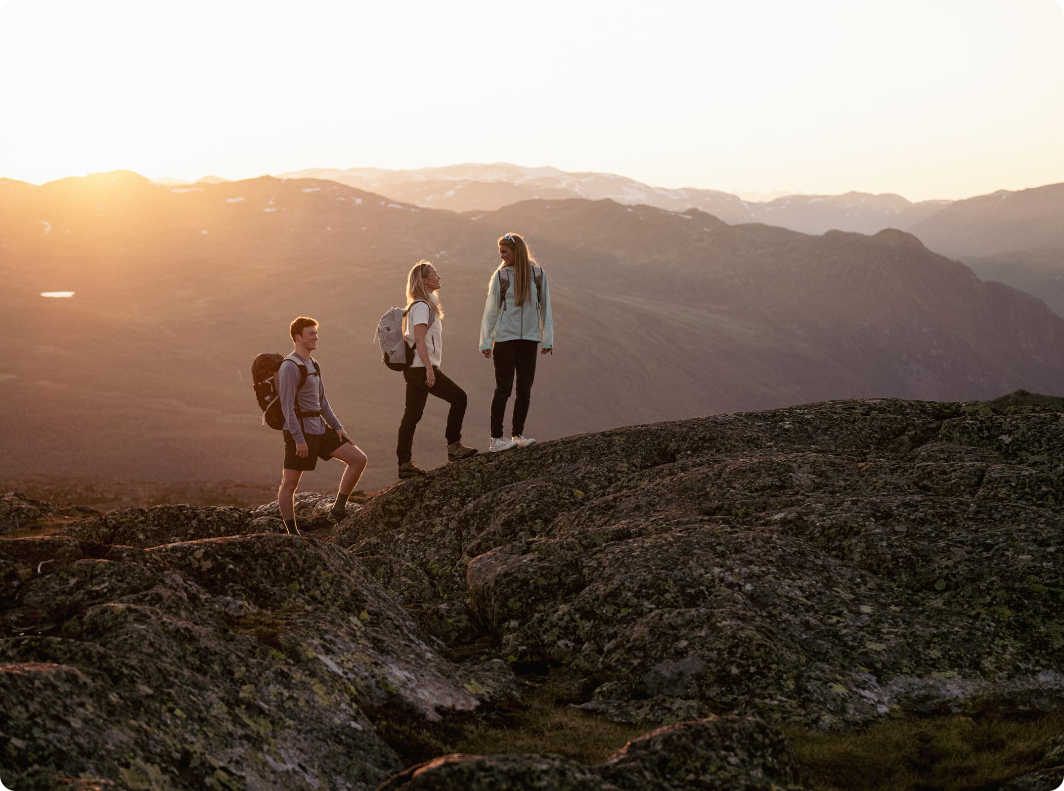 Three hikers with backpacks stand on rocky terrain, enjoying a sunrise view over distant mountains.