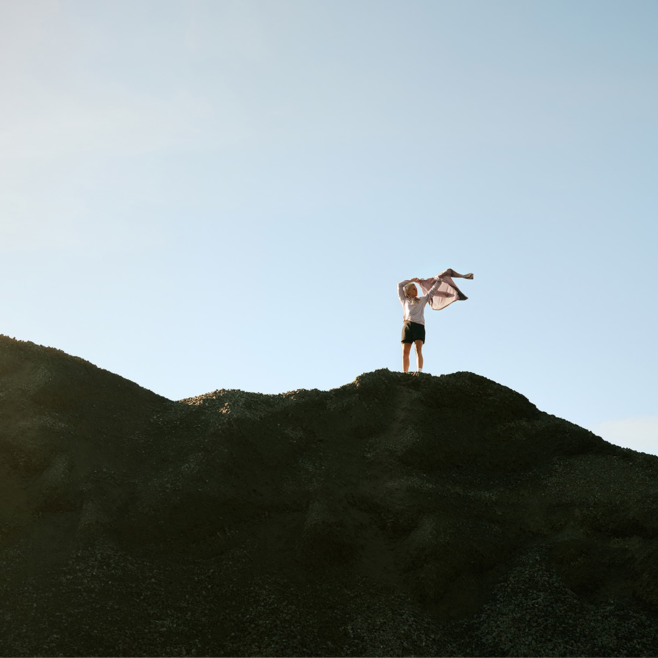 Person standing on a hilltop, holding a jacket, against a clear blue sky.