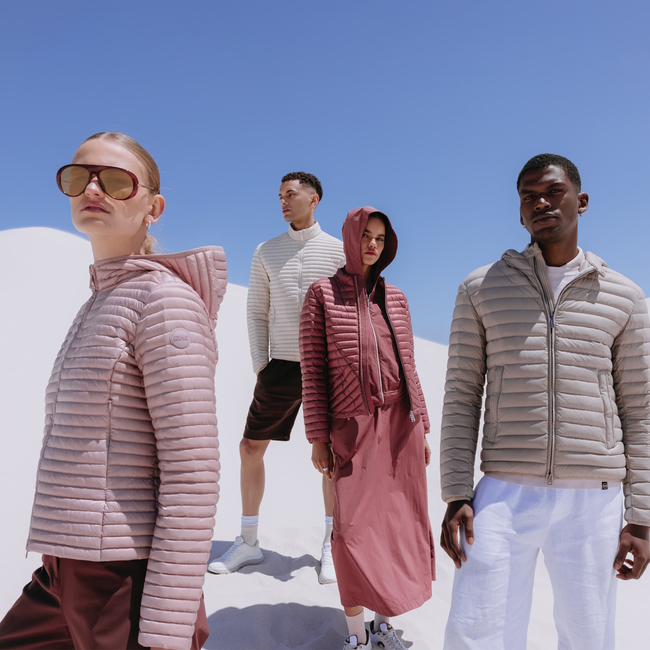 Four people in stylish, puffer jackets stand on white sand under a clear blue sky.