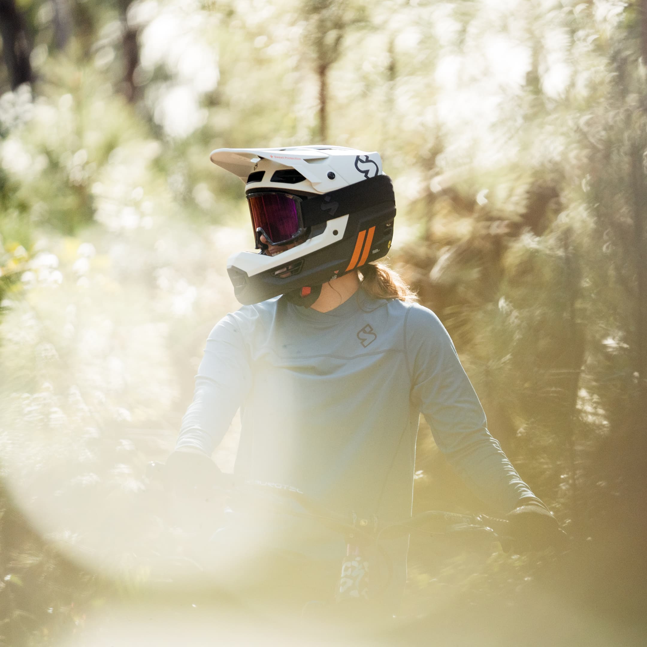A mountain biker wearing a full-face helmet and goggles rides through a sunlit, forested trail.