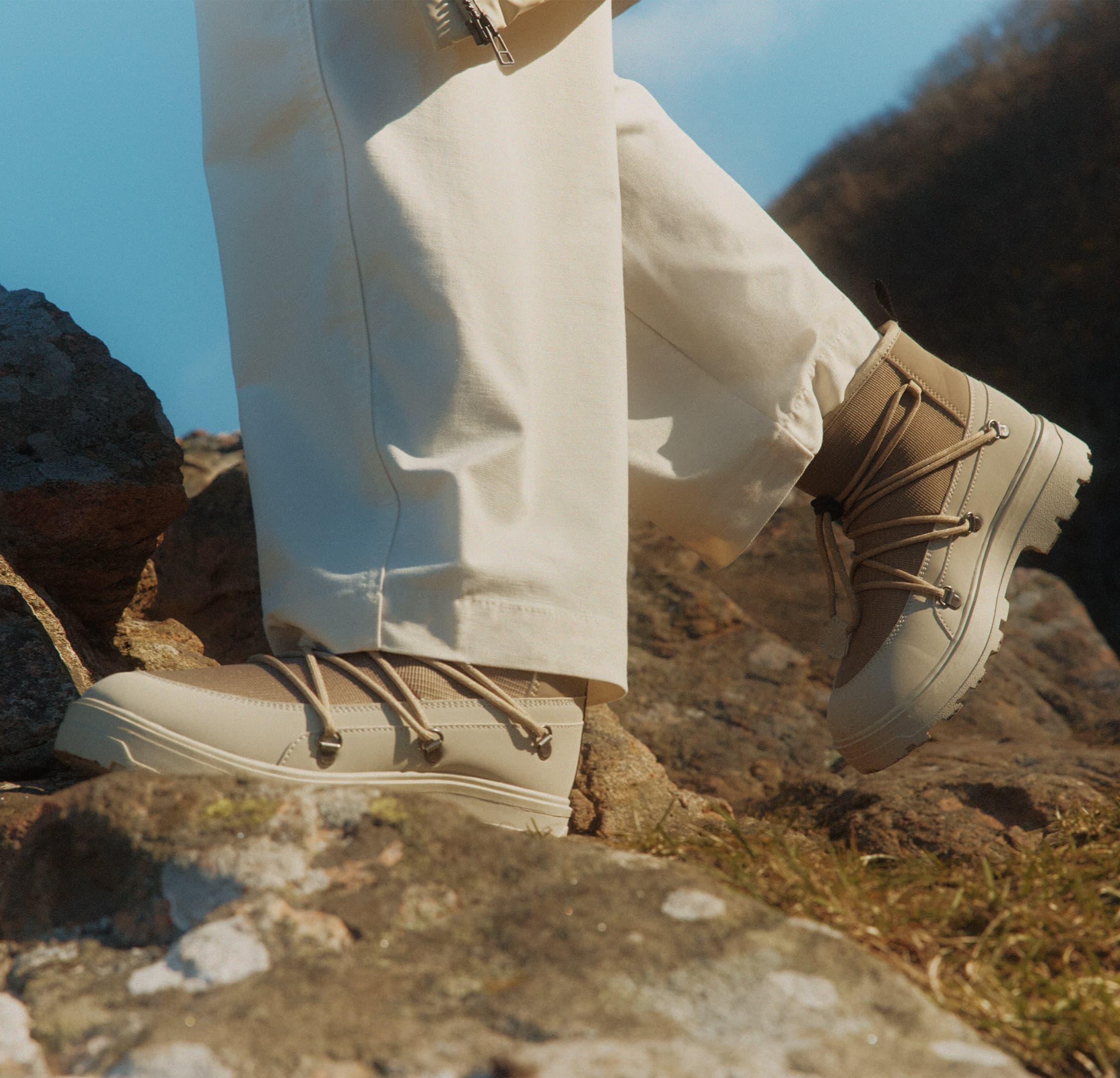 Person wearing beige hiking boots and white pants, standing on rocky terrain with a grassy background under a clear blue sky.