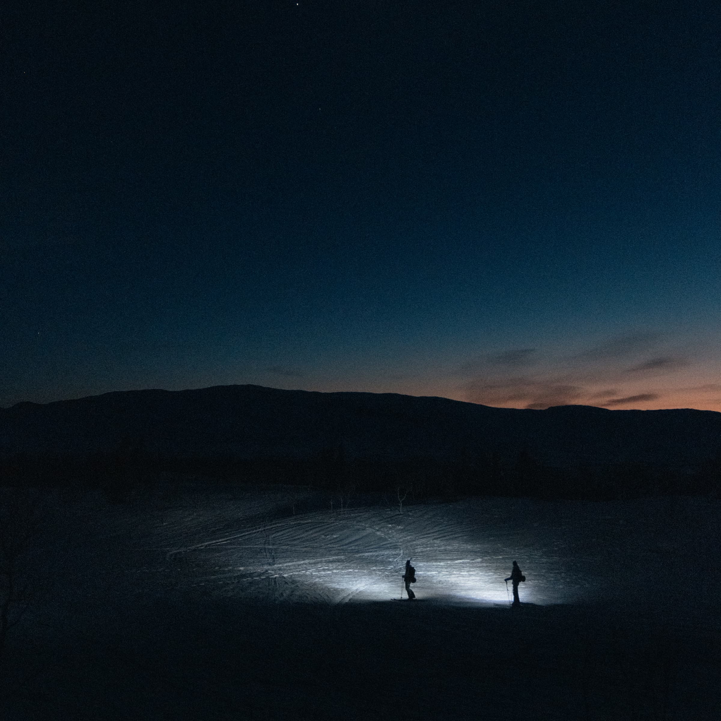 Two people in a snowy landscape, with a dark mountain silhouette and a fading blue and orange sky in the background.