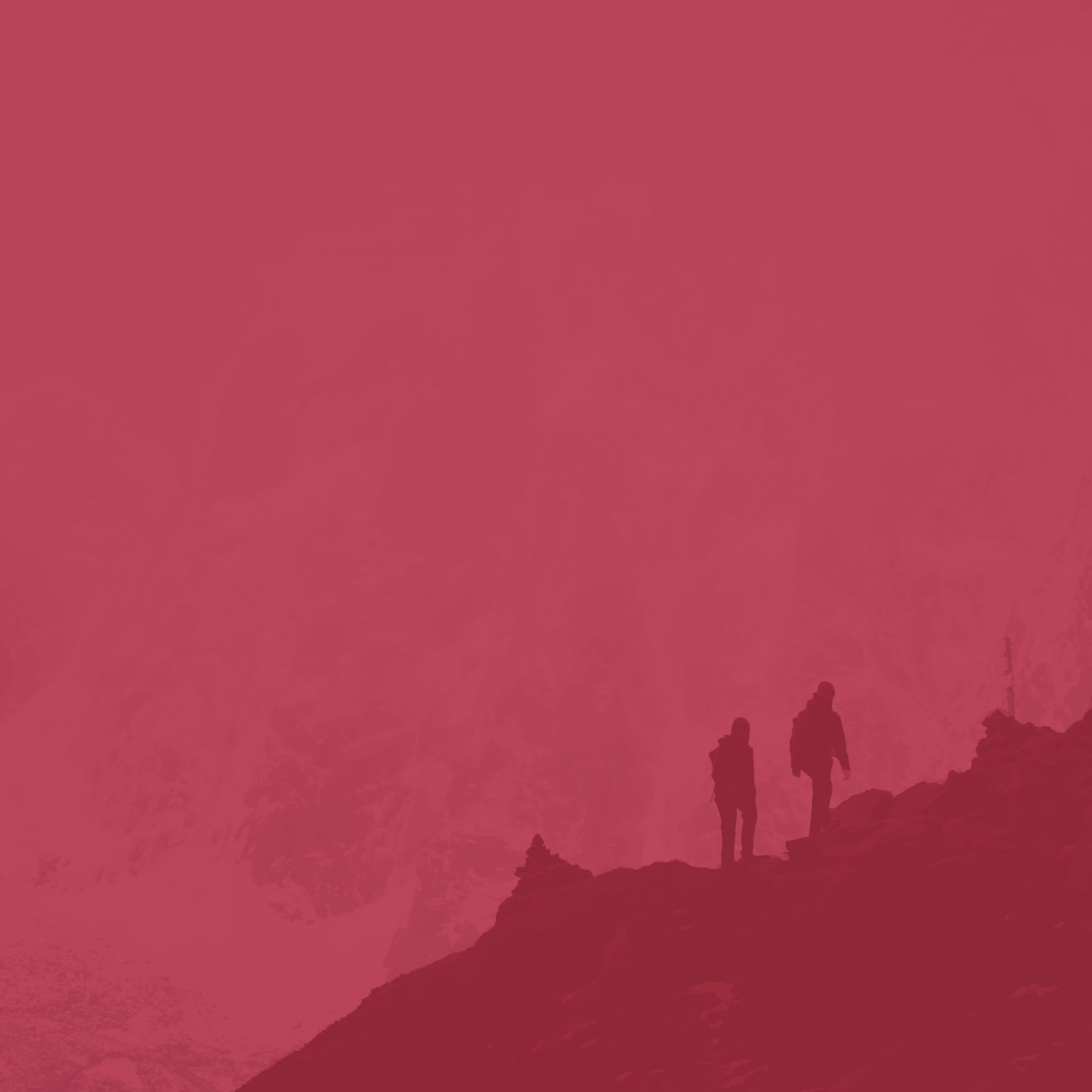 Two silhouetted hikers walks on a rocky ridge, set against a monochromatic red-tinted mountainous background.