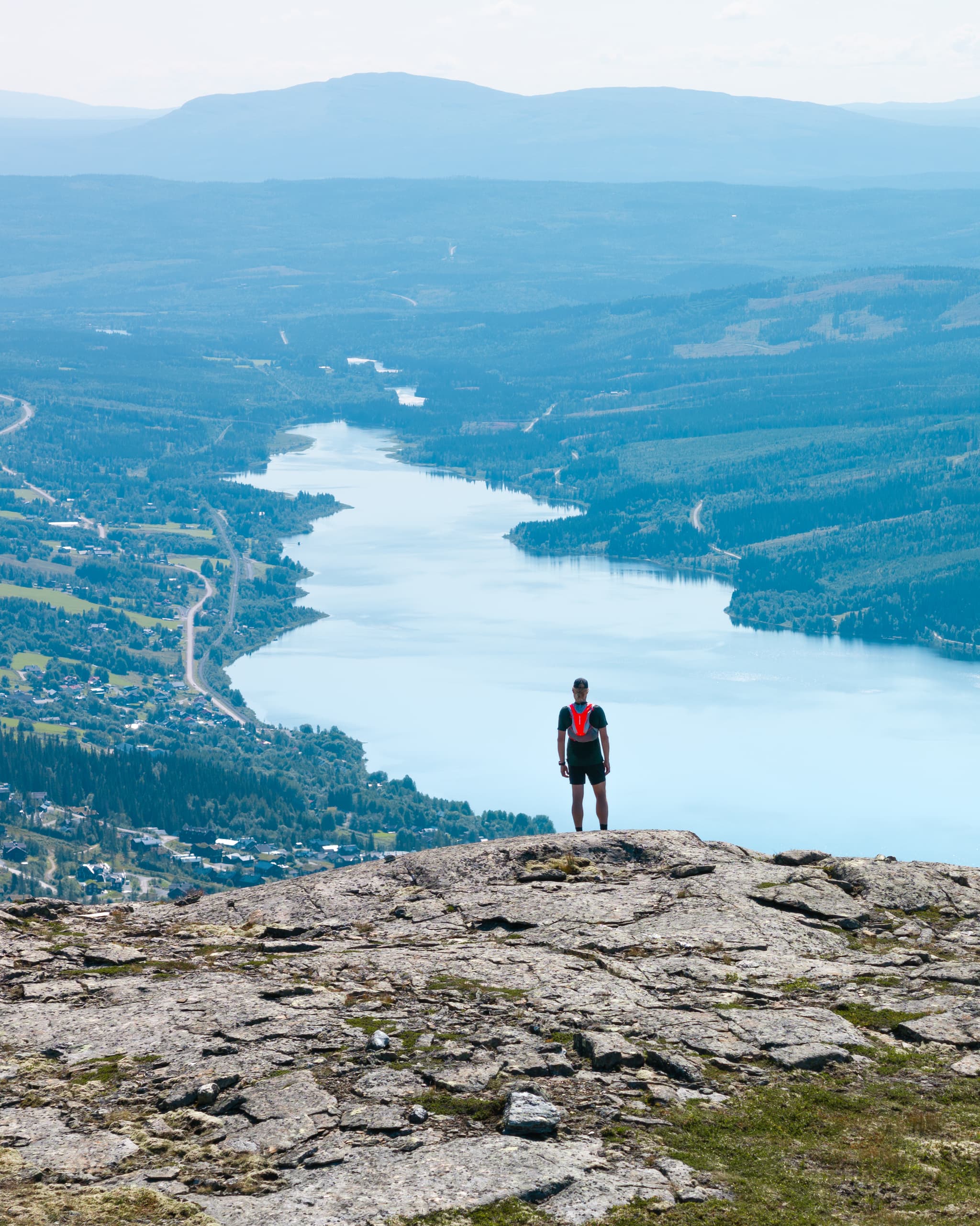 Person standing on a rocky cliff overlooking a winding river and vast landscape with hills and forests in the distance.