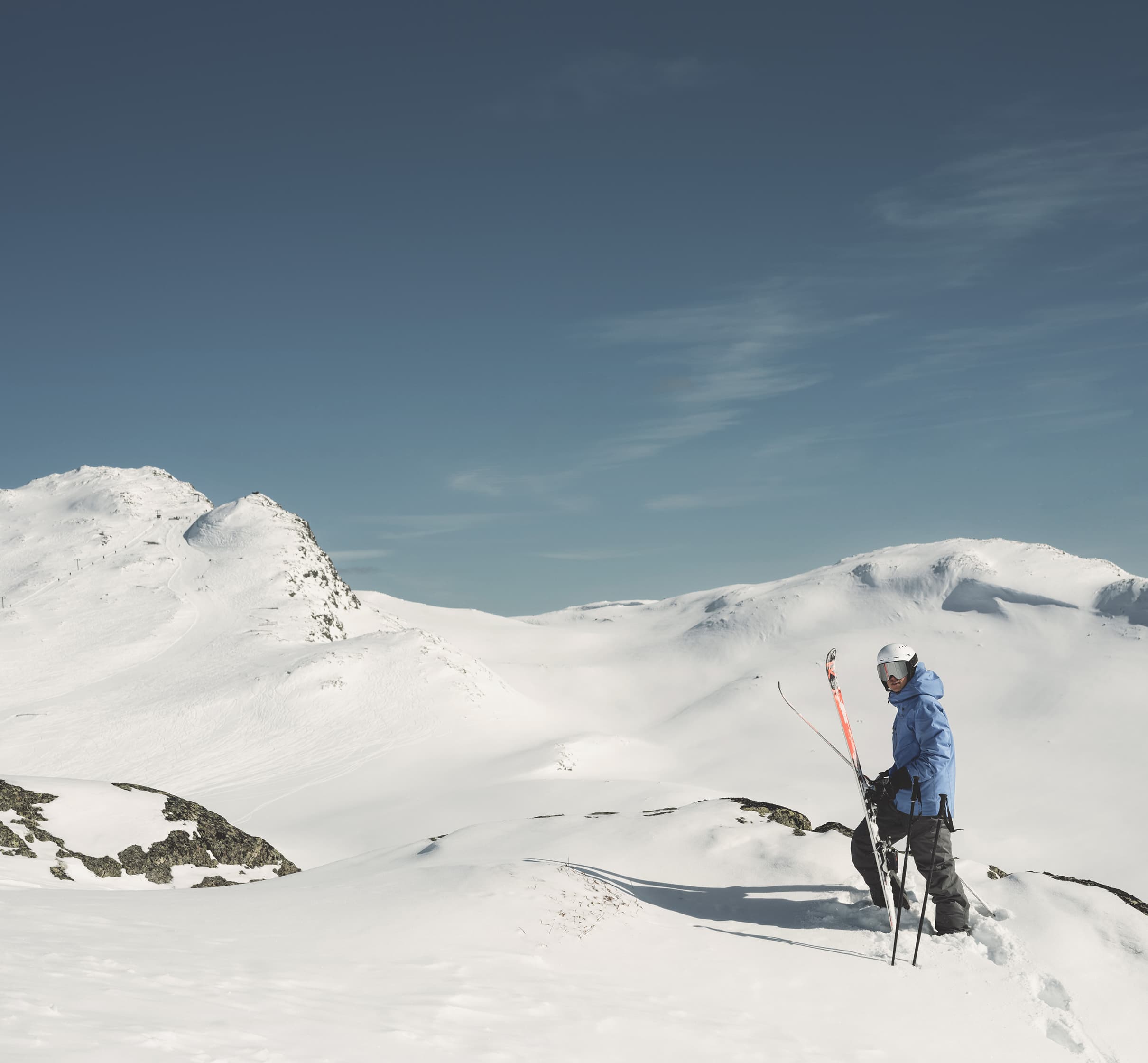 Skier standing on a snowy mountain slope under a clear blue sky, holding ski poles, with distant snow-covered peaks in the background.