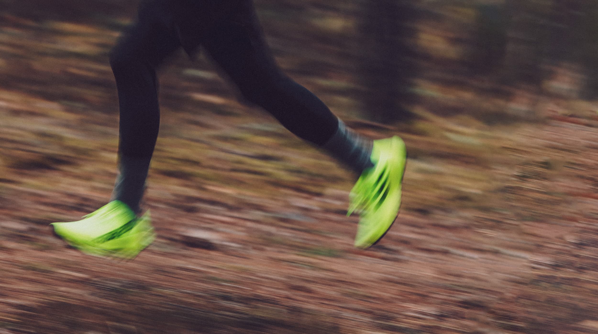 Person running in the forest wearing bright neon green shoes, captured in motion with a blurred background.