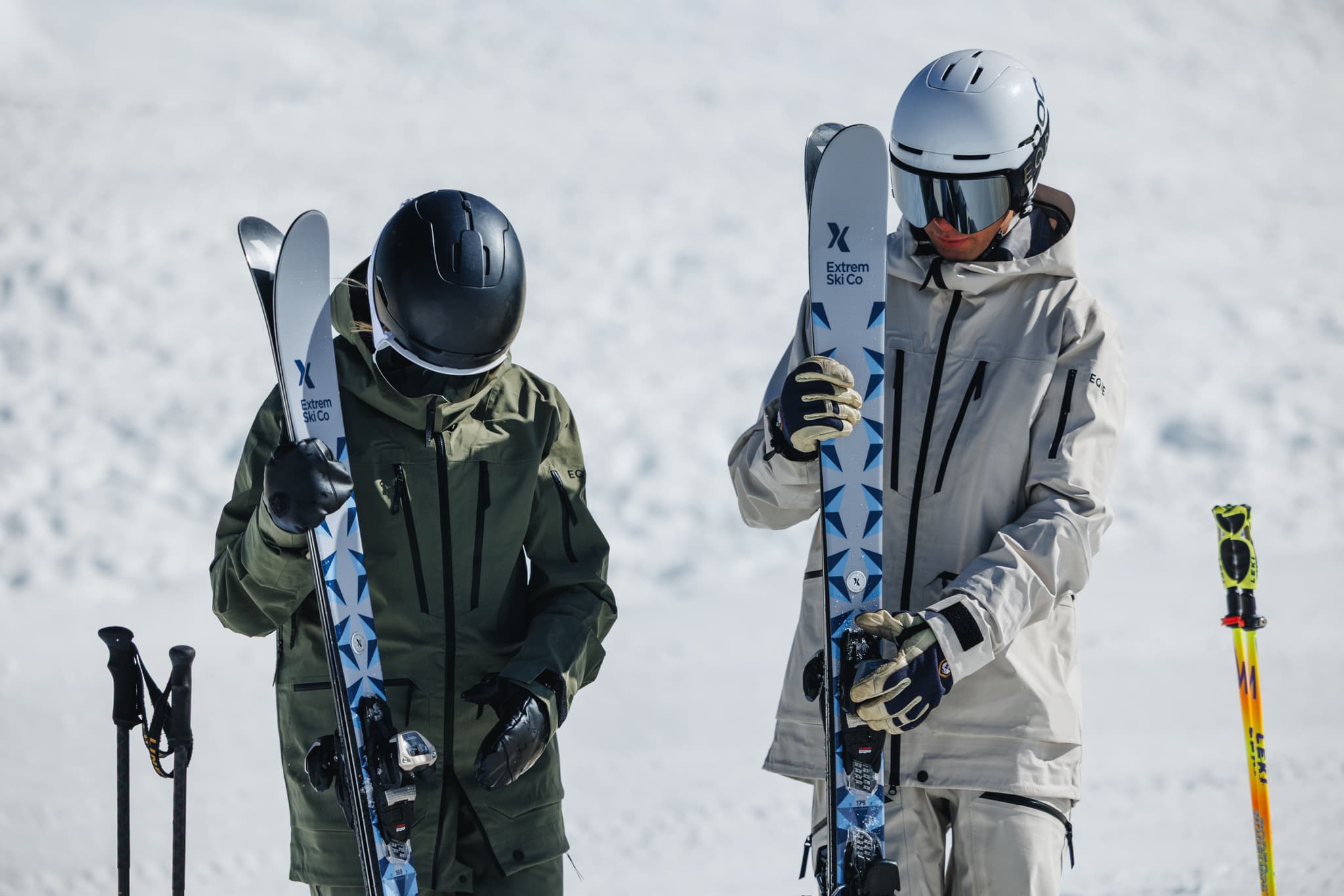 Two skiers in winter gear hold skis vertically on a snowy slope, with ski poles planted in the snow beside them.