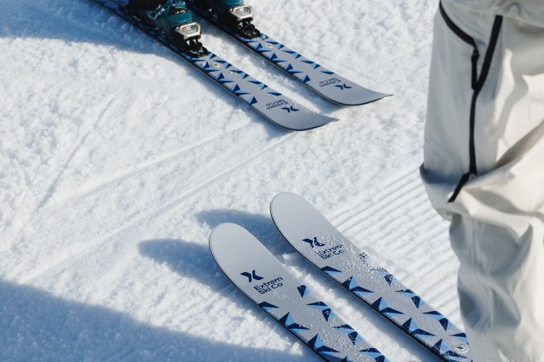 Close-up of two pairs of skis with blue and white designs on a snow-covered surface, with a person in white ski pants nearby.