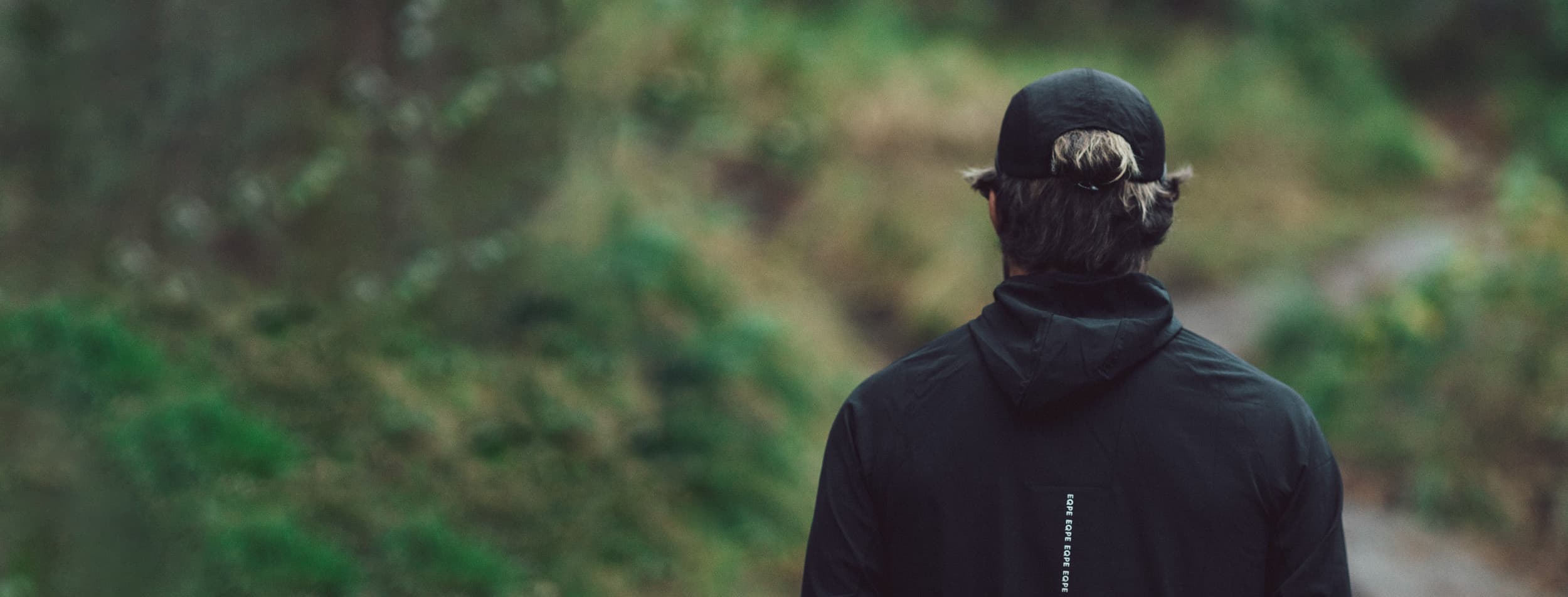 A person in a black jacket and cap walks on a path through a green, forested area, facing away from the camera.