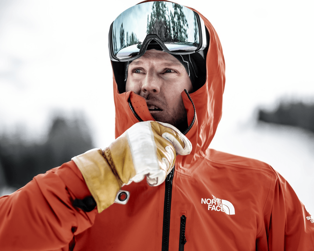 Person in an red jacket and ski goggles, wearing white gloves, stands outdoors in a snowy landscape, looking thoughtfully into the distance.