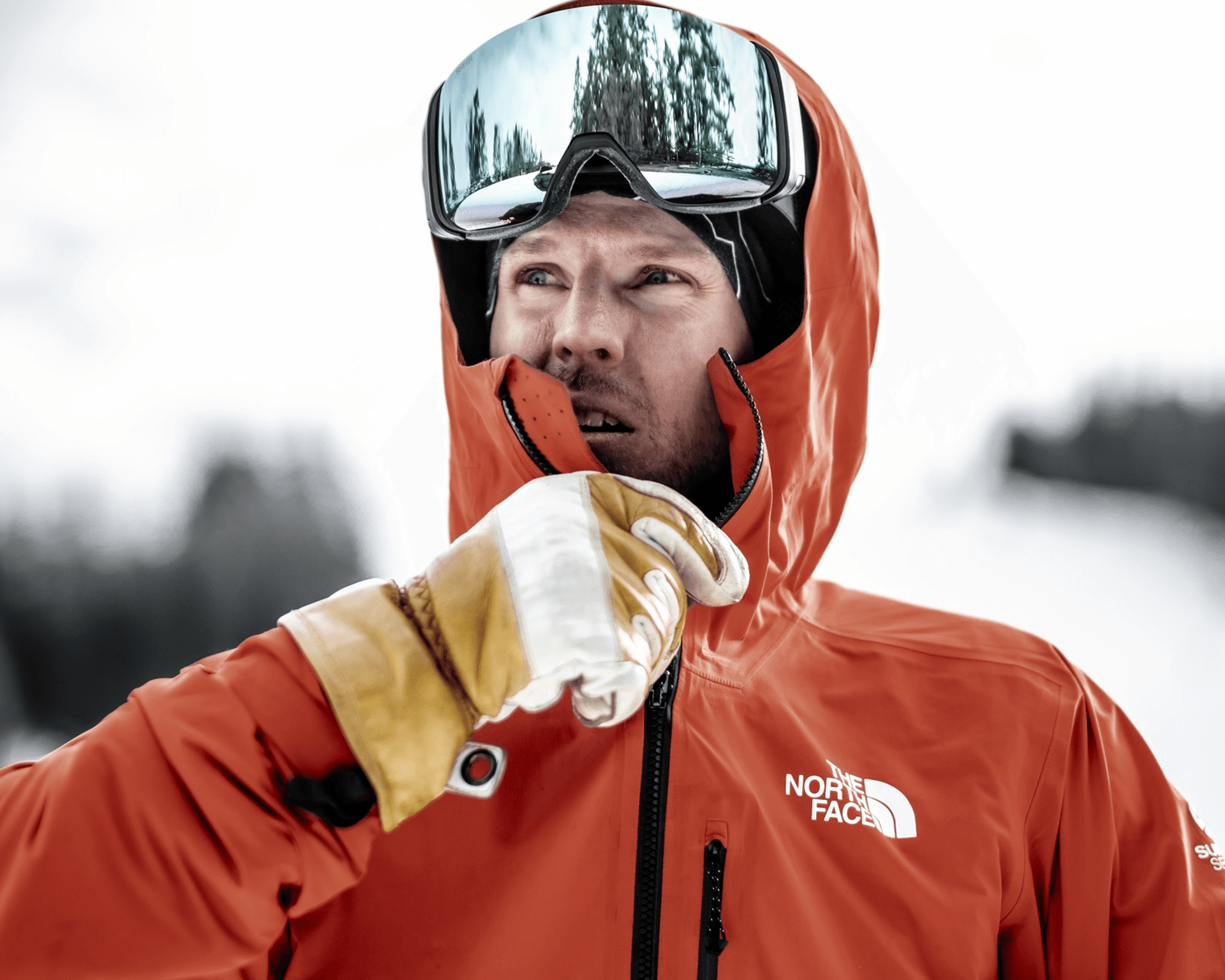 Person in an red jacket and ski goggles, wearing white gloves, stands outdoors in a snowy landscape, looking thoughtfully into the distance.