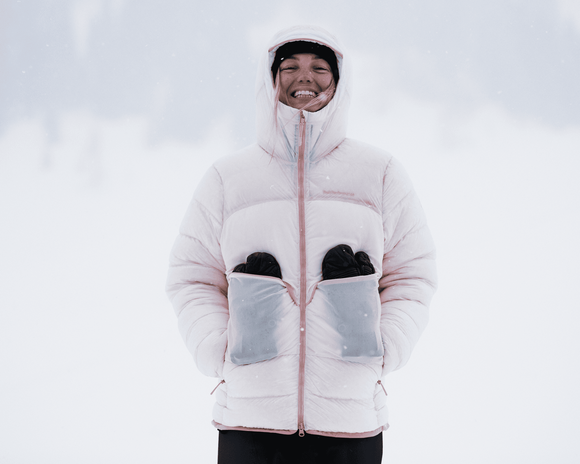Person smiling in a snowy landscape, wearing a pink winter jacket with gloves tucked into front pockets.