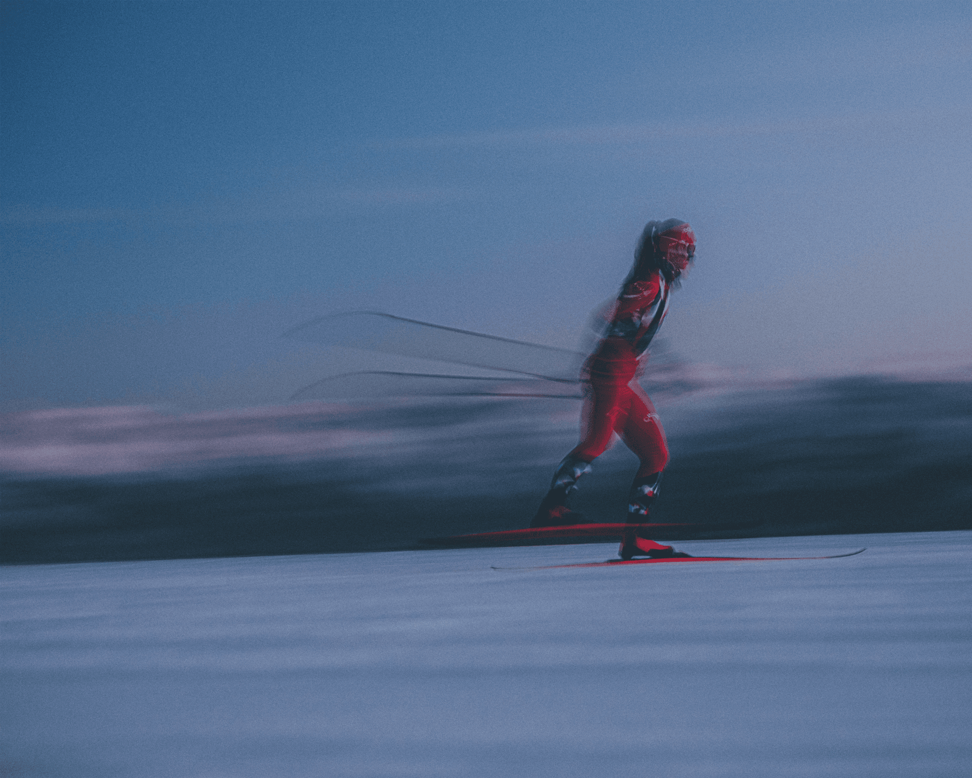 A cross-country skier in motion, wearing red gear, glides swiftly on snow against a blurred, twilight sky backdrop.