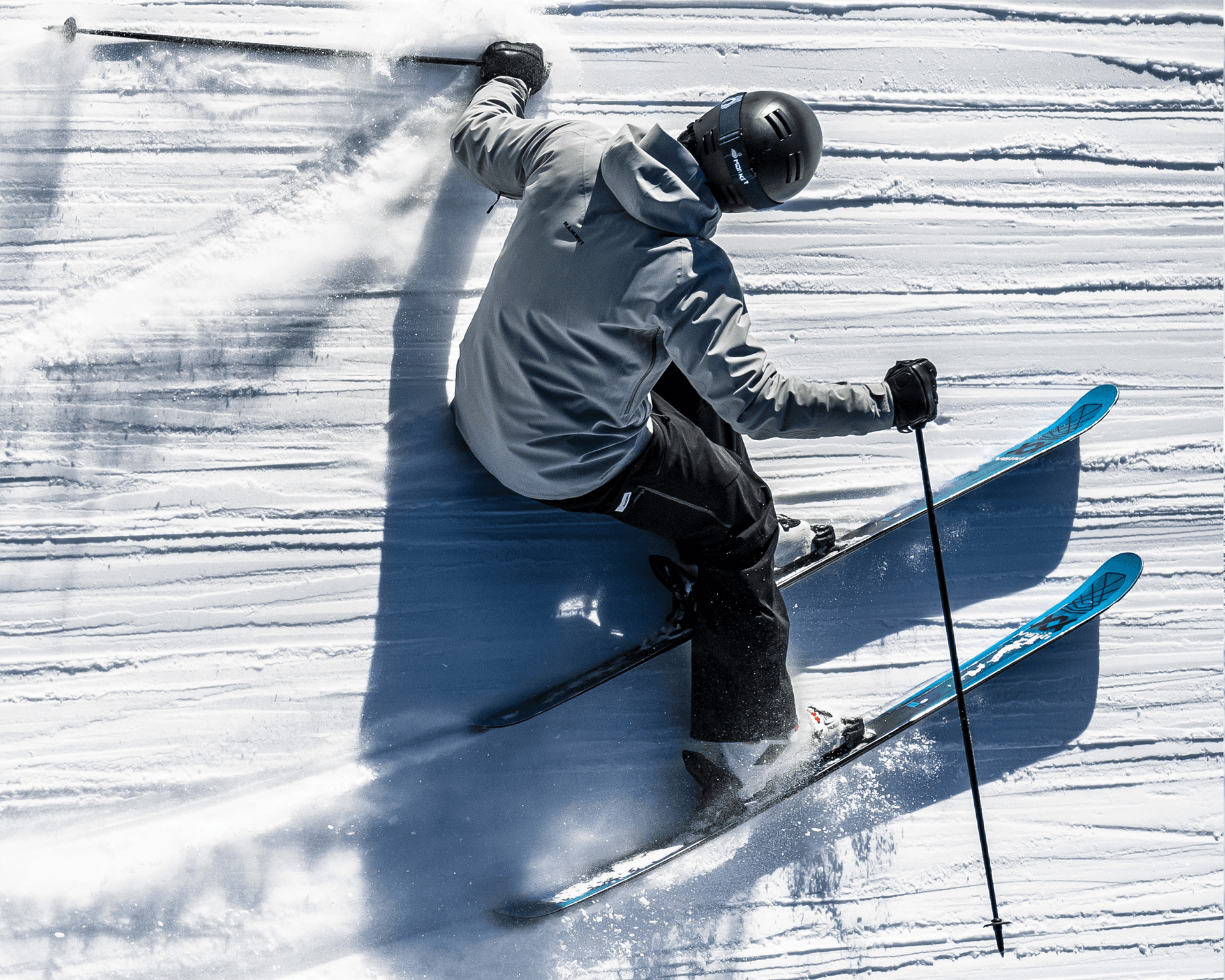 Skier in a gray jacket and black pants making a sharp turn on a groomed snowy slope, creating a spray of snow.