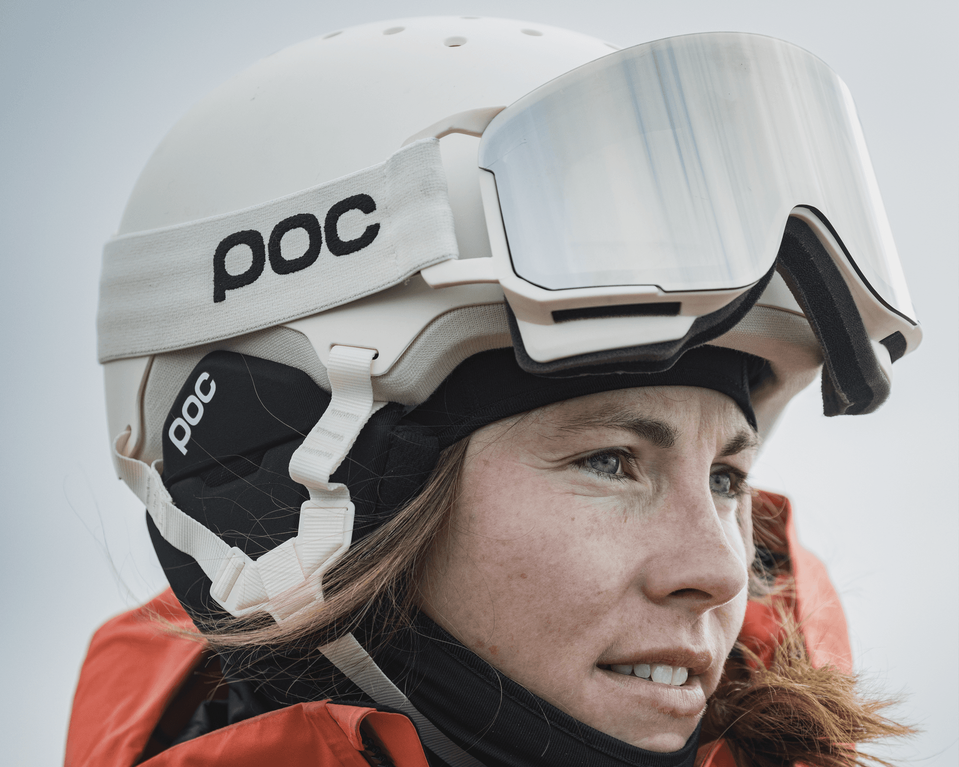 Person in a white ski helmet and goggles, wearing an orange jacket, looking into the distance against a light sky.