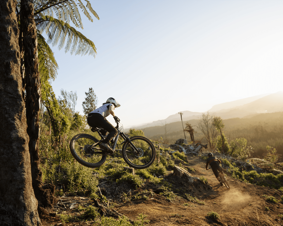 Two mountain bikers riding on a dirt trail; one is airborne, surrounded by lush greenery and a distant view of hills.
