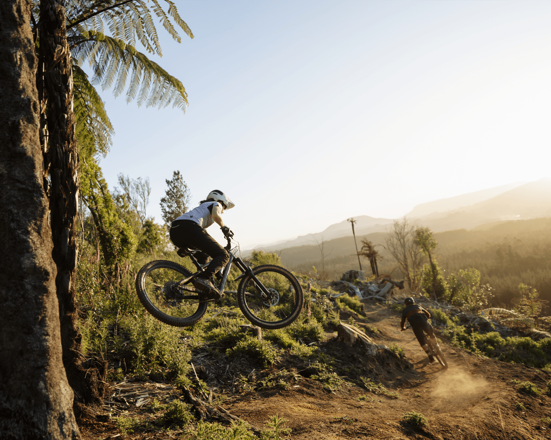 Two mountain bikers riding on a dirt trail; one is airborne, surrounded by lush greenery and a distant view of hills.