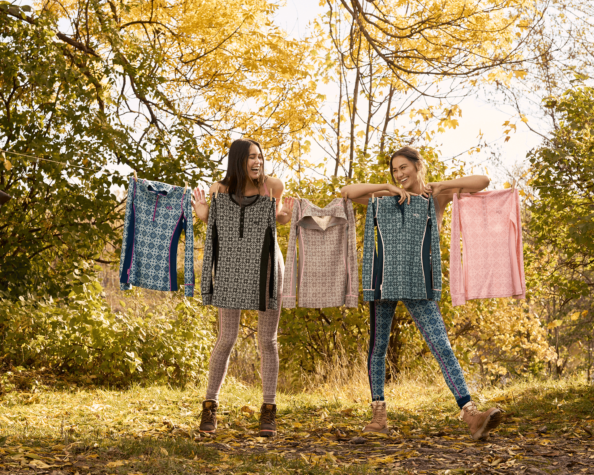 Two women standing outdoors on a sunny day, smiling and holding up patterned base layer tops on a clothesline. They are surrounded by trees with golden-yellow leaves and are dressed in colorful base layers and boots.