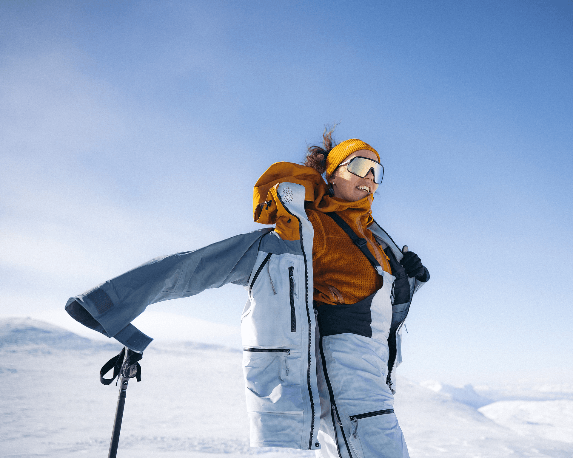 Person in winter gear skiing on a snowy mountain, wearing sport eyewear and a yellow headband, against a clear blue sky.