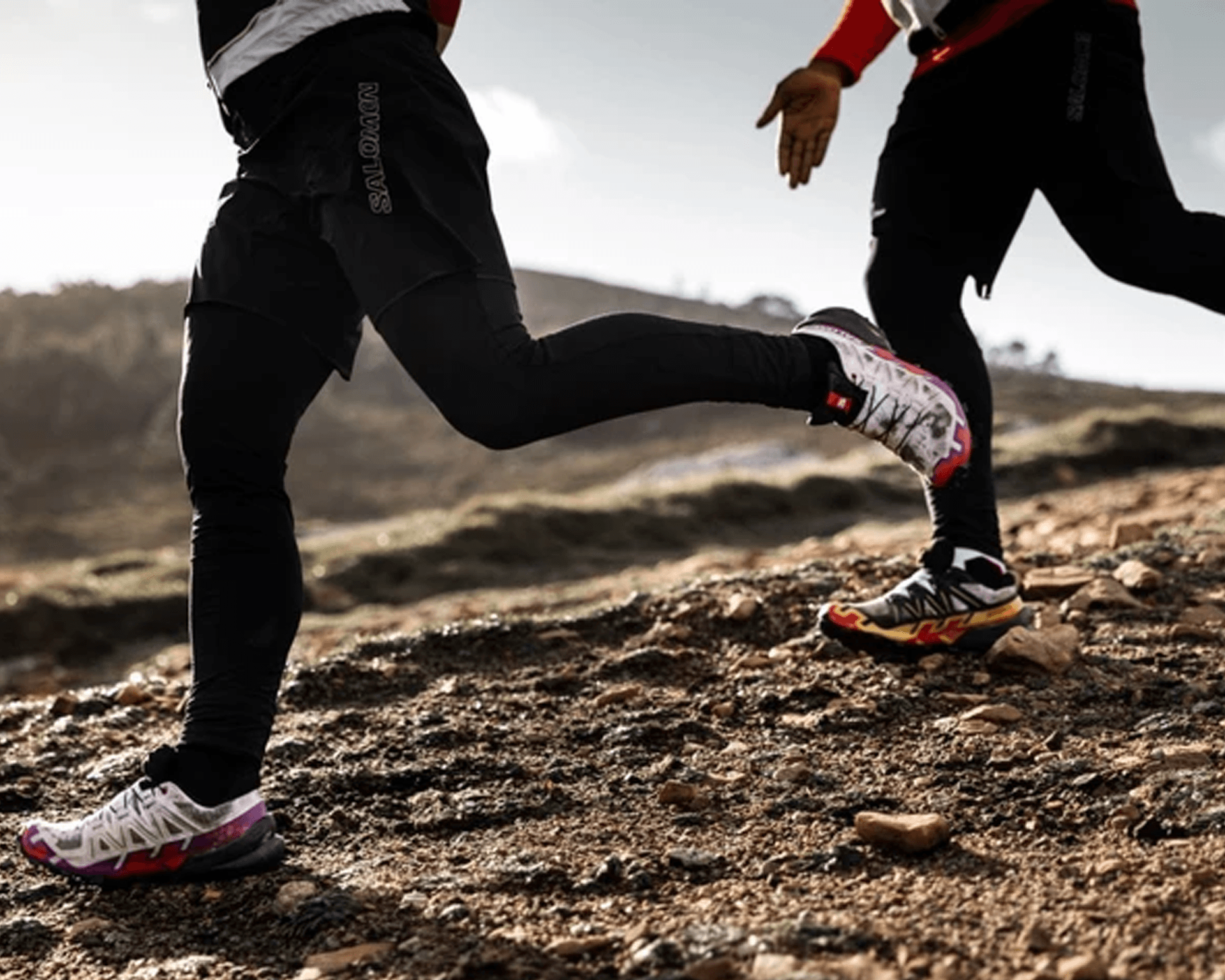 Two people trail running on a rocky path, wearing black leggings and colorful running shoes, with a hilly landscape in the background.