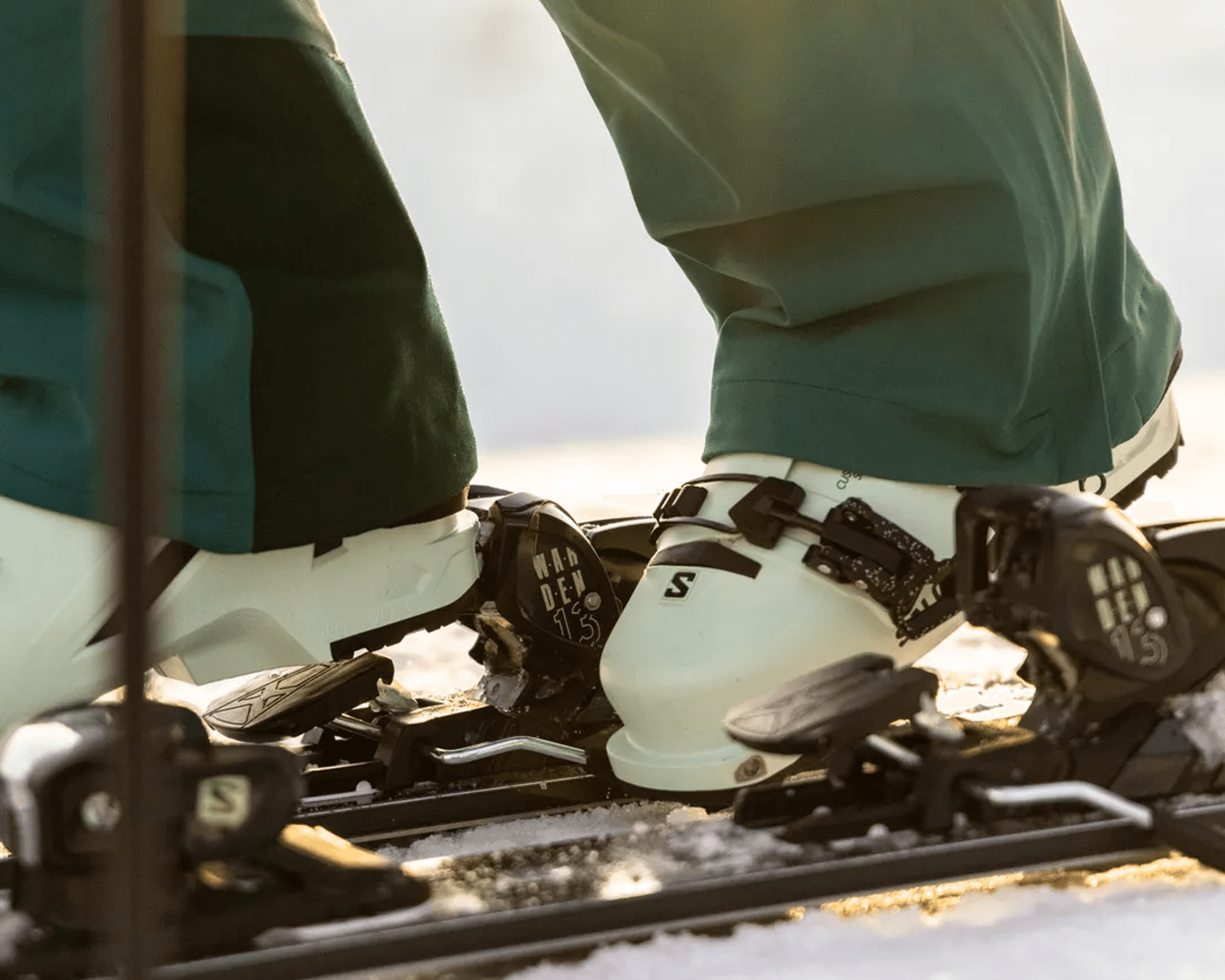 Close-up of a person wearing green ski pants and white ski boots clipped into black ski bindings on a snowy surface.