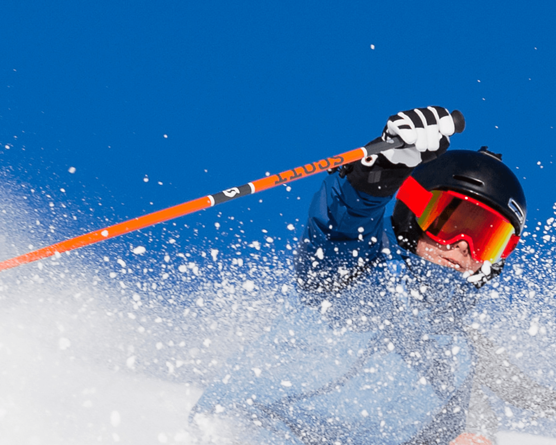 Young skier in blue jacket and red goggles skiing through fresh powder against a clear blue sky.