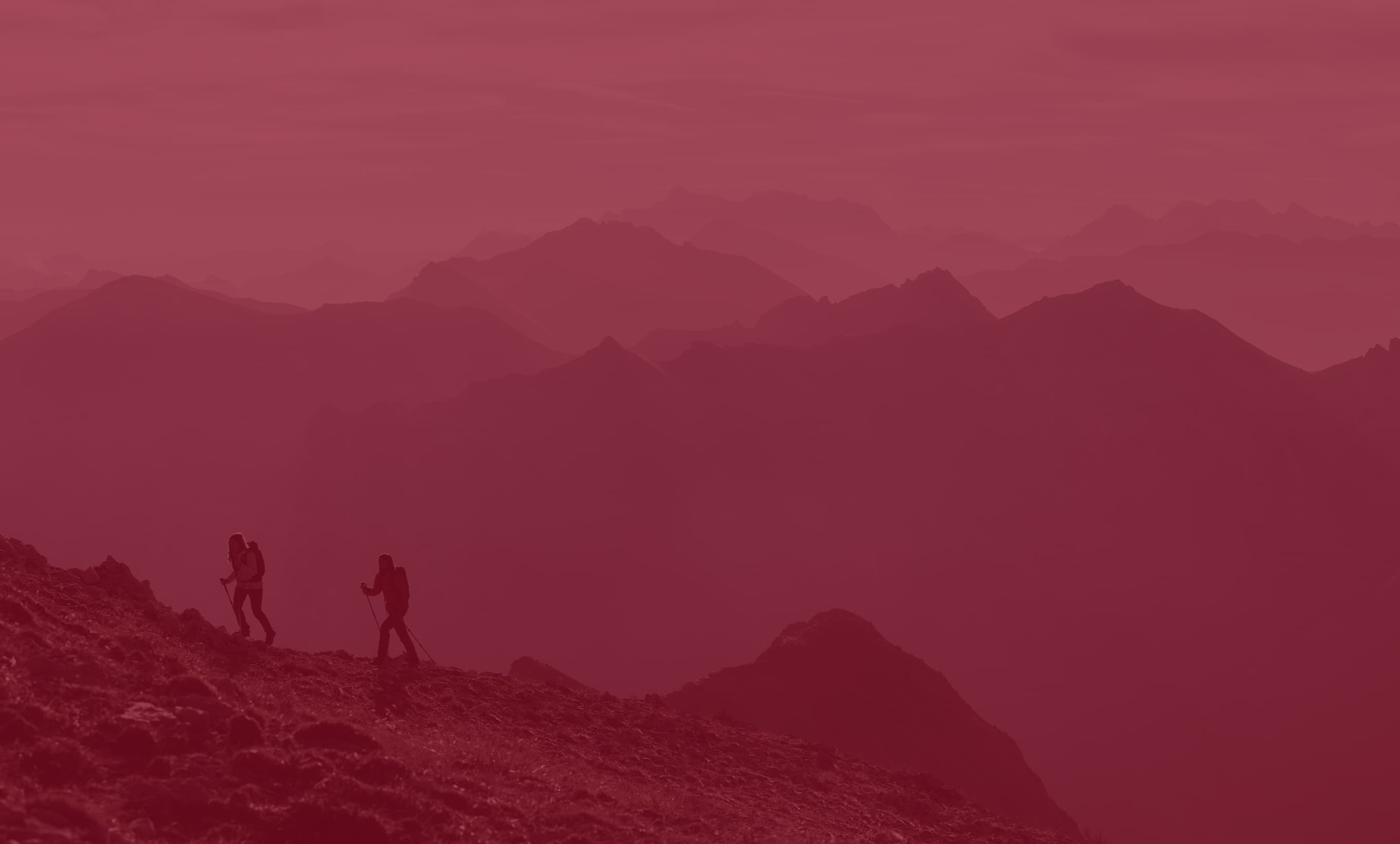 Two hikers traverse a mountain ridge, silhouetted against a hazy, red-tinted backdrop of distant peaks.