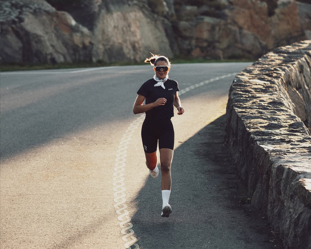 A woman wearing sunglasses and athletic gear runs along a curving road bordered by a stone wall with rocky hills in the background.
