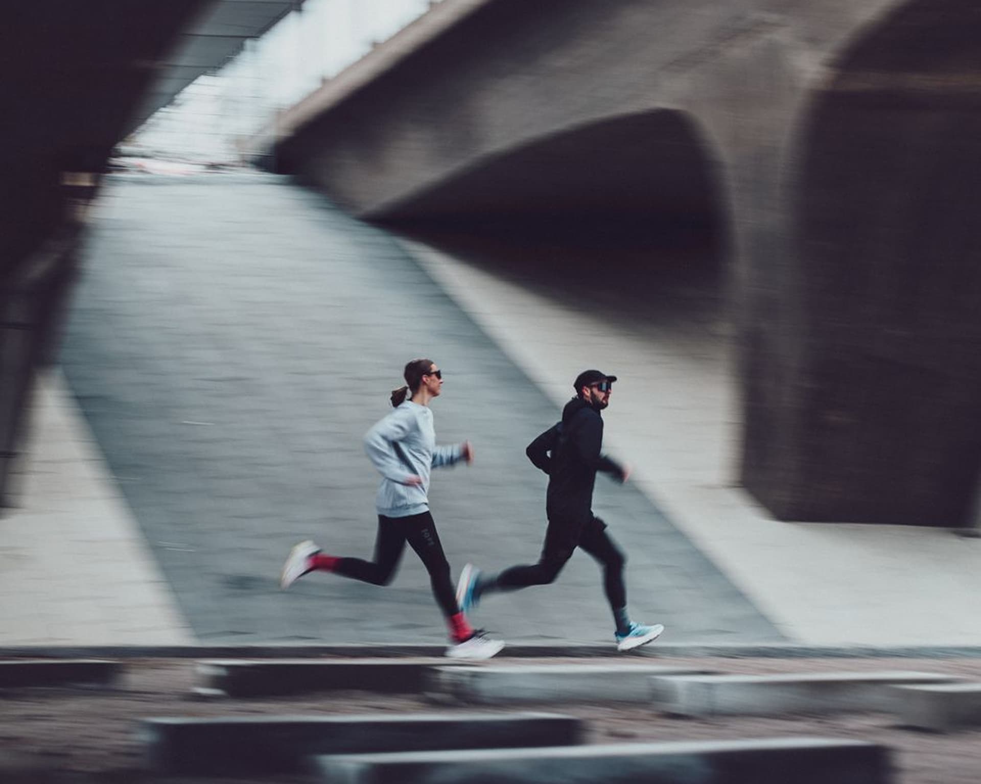 Two people in athletic gear runs under a concrete bridge, their motion blurred to convey speed