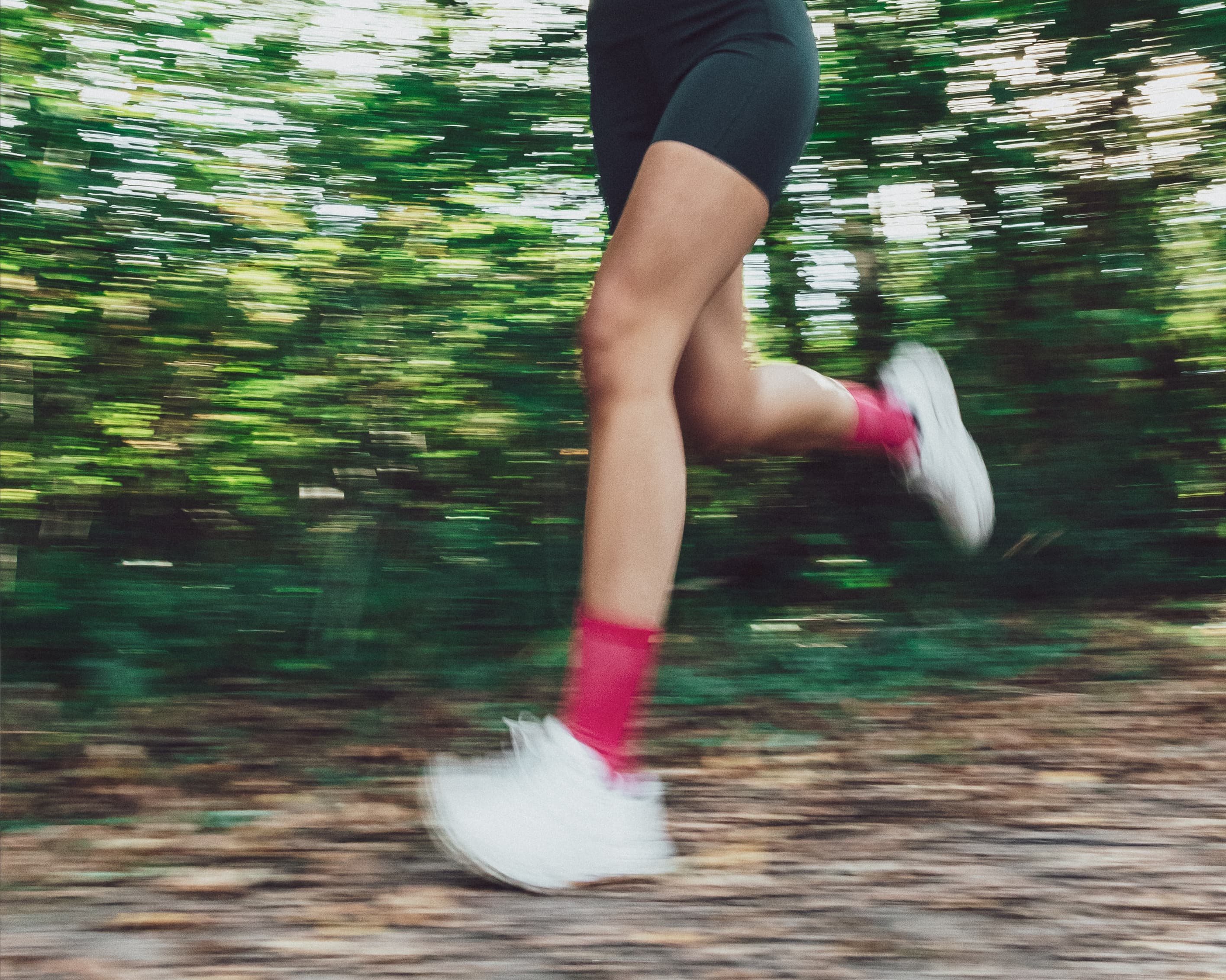Person running outdoors with blurred green background, wearing black shorts, white shoes, and bright pink socks.