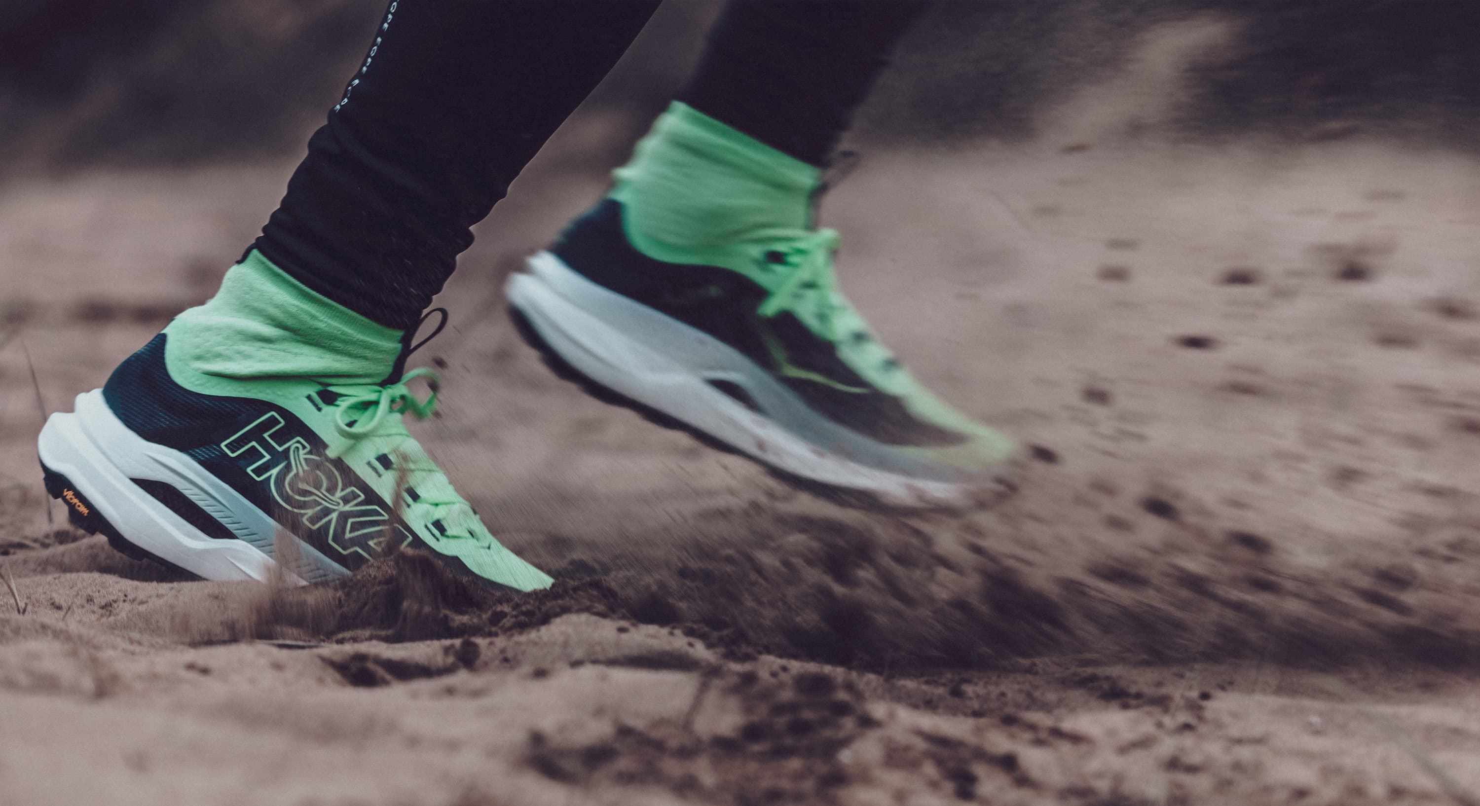 Close-up of a person running on sandy terrain, wearing neon green and black HOKA sneakers with matching socks, creating sand splashes.