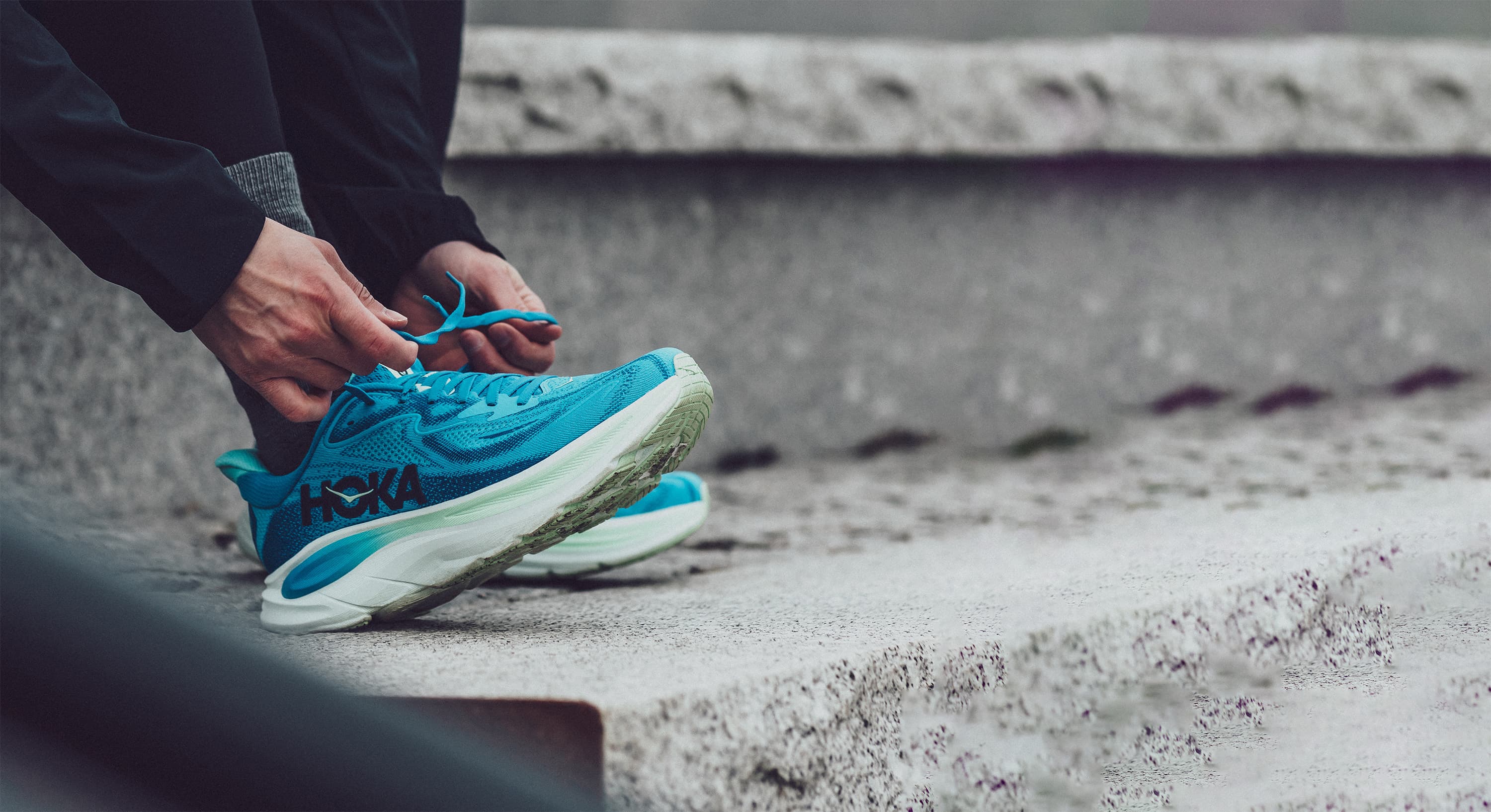 Person tying laces on blue running shoes while sitting on stone steps, wearing dark pants.