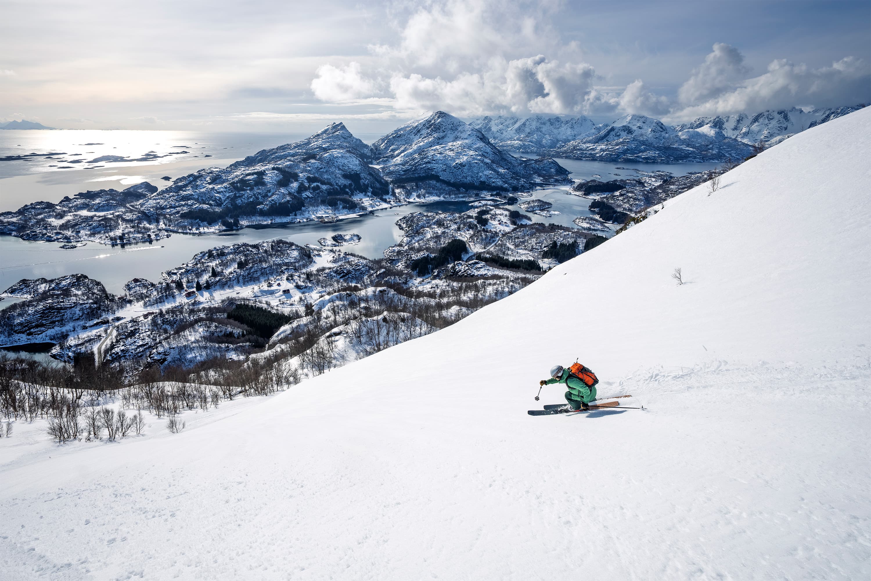 Skier in green gear descends a snowy mountain slope with scenic ocean and mountain views under a partly cloudy sky.