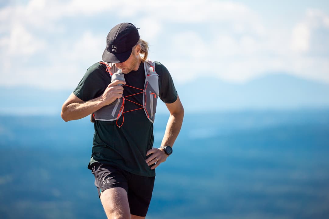 Man in a cap and hydration vest stands on a mountain trail with a scenic view of blue skies and distant hills.
