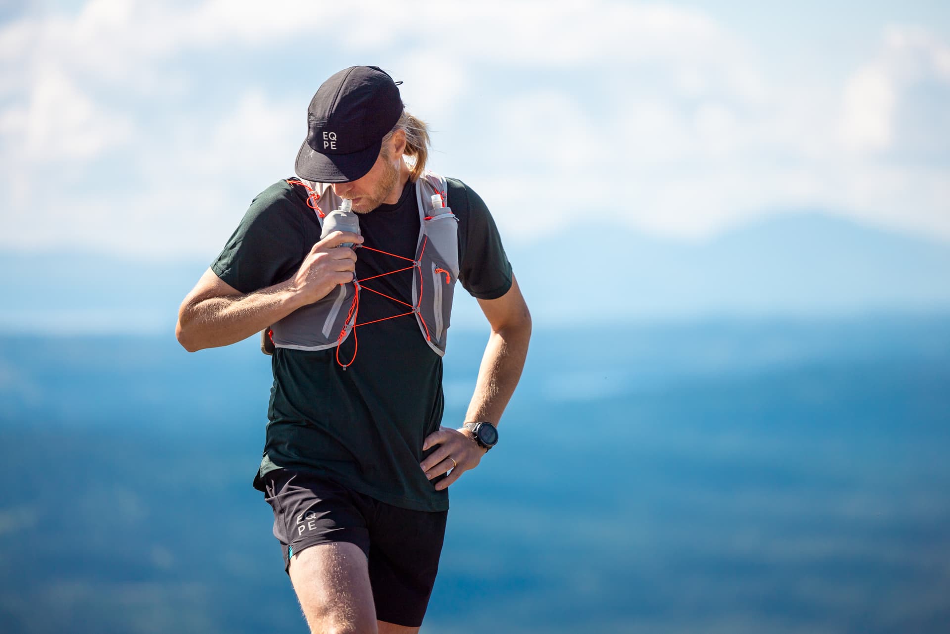 Man in a cap and hydration vest stands on a mountain trail with a scenic view of blue skies and distant hills.