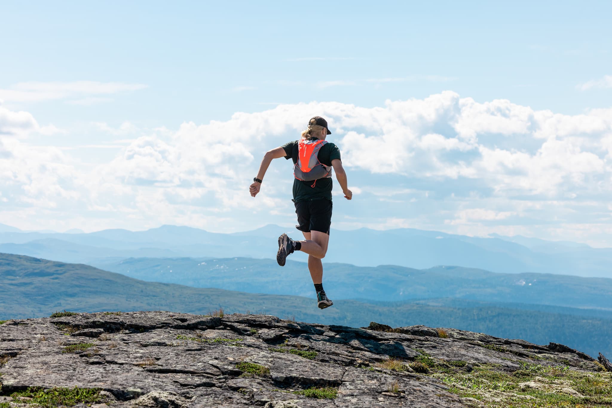 Person running on a rocky mountain trail with a colorful running vest, under a clear blue sky and distant mountain range.